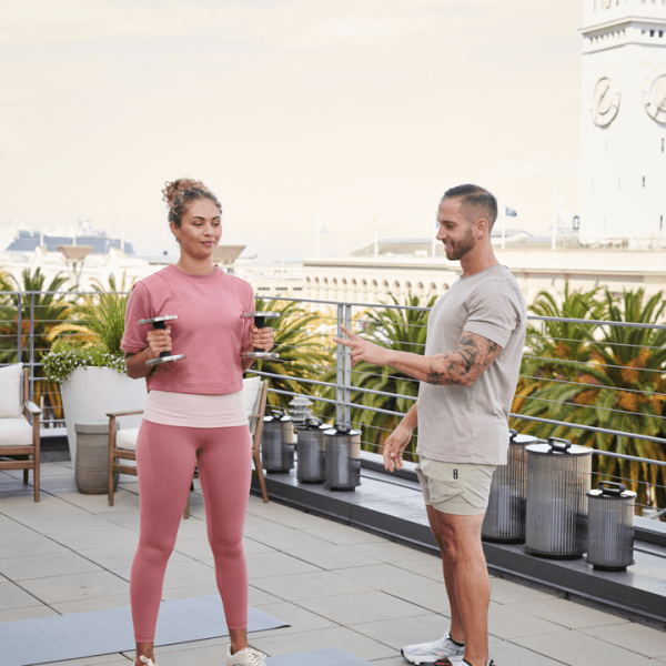 A male private trainer instructs a female client holding free weights, on the rooftop of 1 Hotels SFO