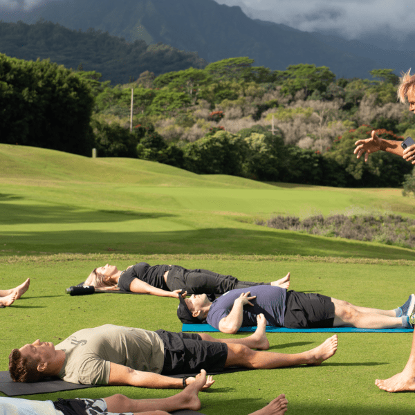 Group of people laying on yoga mats outside while an instructor guides them